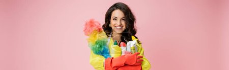 cheerful brunette housewife with colorful feather duster and detergents looking at camera isolated on pink, banner