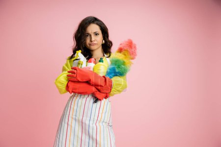 frowning housewife in striped apron and red rubber gloves holding pile of cleaning supplies isolated on pink