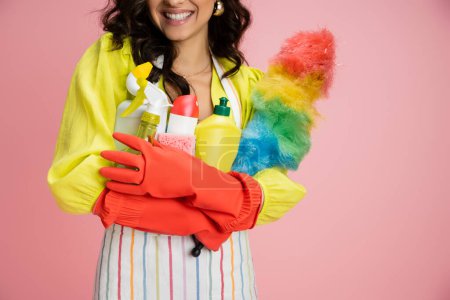 vista recortada de ama de casa sonriente en guantes de goma roja sosteniendo un montón de artículos de limpieza aislados en rosa
