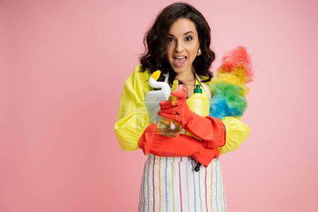 astonished housewife with cleaning supplies having fun while spraying from bottle at camera isolated on pink