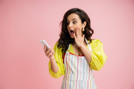 Foto de Sorprendido ama de casa en delantal rayado tocando la cara mientras mira el teléfono móvil aislado en rosa - Imagen libre de derechos