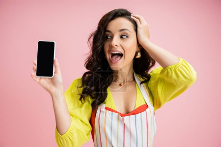 Foto de Mujer asombrada en blusa amarilla y delantal rayado tocando la cabeza mientras sostiene el teléfono celular con pantalla en blanco aislado en rosa - Imagen libre de derechos