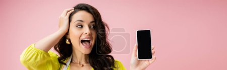 amazed housewife touching wavy hair while looking at cellphone with blank screen isolated on pink, banner