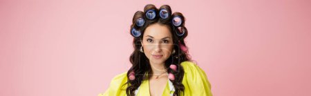 portrait of brunette housewife in hair curlers and bright yellow blouse looking at camera isolated on pink, banner