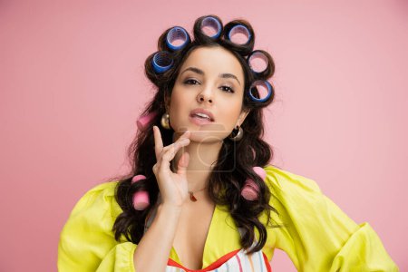 sensual brunette housewife in yellow blouse and hair curlers touching chin and looking at camera isolated on pink