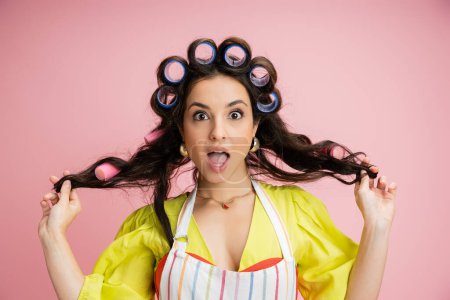 surprised housewife in hair curlers and yellow blouse holding hair and looking at camera isolated on pink