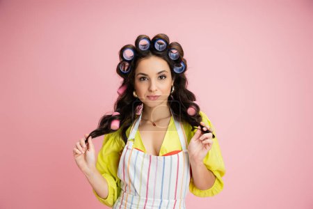 brunette housewife in hair curlers and striped apron looking at camera isolated on pink