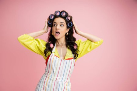 amazed and curious housewife in striped apron adjusting hair curlers and looking away isolated on pink