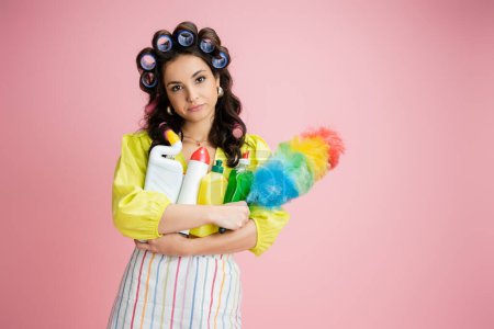 bored housewife with hair curlers and different cleaning supplies looking at camera isolated on pink