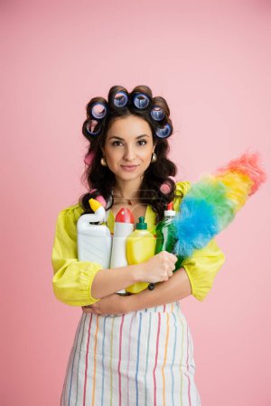 positive housewife in hair curlers holding cleaning supplies and colorful dust brush isolated on pink