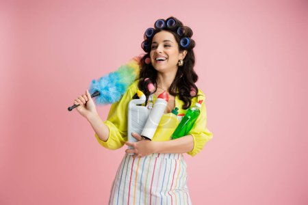 cheerful housewife in hair curlers holding colorful feather duster and cleaning supplies while looking away isolated on pink