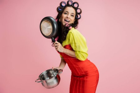 happy housewife in red corset dress holding kitchenware while posing in hair curlers isolated on pink