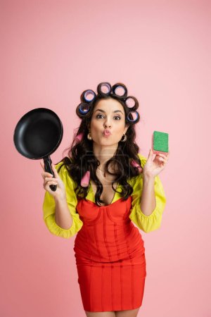 young housewife in hair curlers pouting lips while holding frying pan and sponge isolated on pink