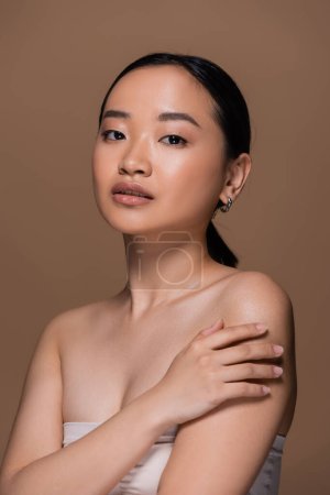 Foto de Young asian woman in top touching shoulder and looking at camera isolated on brown - Imagen libre de derechos