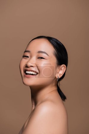 Positive asian model with naked shoulder looking at camera isolated on brown 