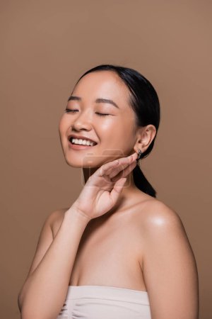 Happy asian woman in top closing eyes and touching face isolated on brown 