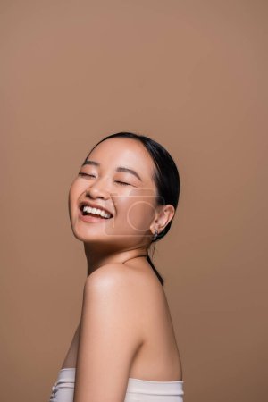 Joyful asian woman in top closing eyes while posing isolated on brown 