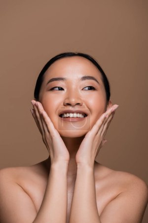 Cheerful asian woman touching face and looking away isolated on brown 