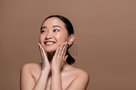 Photo for Cheerful asian model with naked shoulders touching cheeks isolated on brown - Royalty Free Image