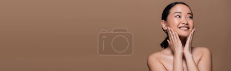 Foto de Positive asian woman with makeup and naked shoulders touching cheeks isolated on brown, banner - Imagen libre de derechos
