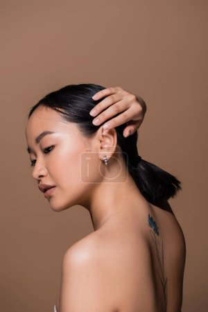 Tattooed asian woman touching hair isolated on brown 