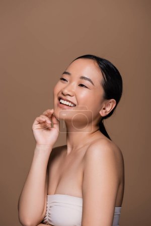 Positive asian woman in top touching face and looking away isolated on brown 