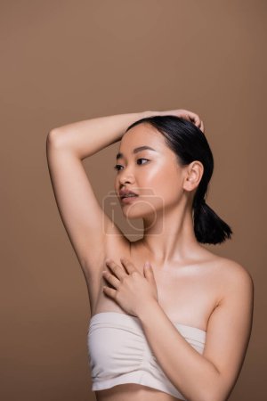 Young asian model in beige top with naked shoulders posing isolated on brown 