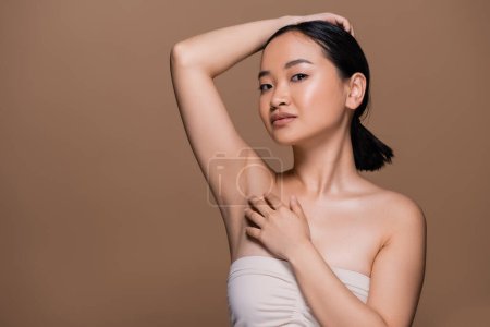 Téléchargez les photos : Pretty asian woman with naked shoulders touching fair and looking at camera isolated on brown - en image libre de droit