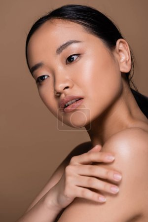 Young asian woman with natural makeup touching naked shoulder isolated on brown 