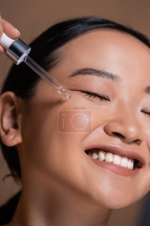 Close up view of smiling asian woman applying cosmetic serum on face isolated on brown 