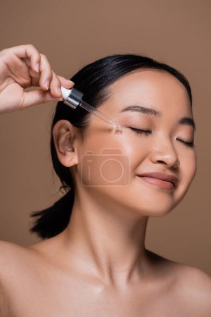 Joyful asian woman with dropper applying cosmetic serum on cheek isolated on brown 