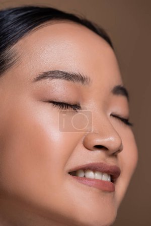 Close up view of smiling asian model with natural makeup isolated on brown 