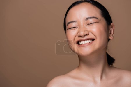 Foto de Happy asian woman with naked shoulders and closed eyes standing isolated on brown - Imagen libre de derechos
