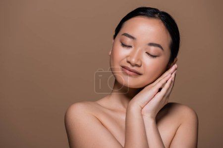 Young brunette asian woman with naked shoulders holding hands near cheek isolated on brown 