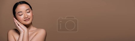 Foto de Pretty asian woman with naked shoulders holding hands near cheek isolated on brown, banner - Imagen libre de derechos