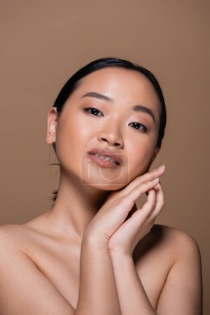 Portrait of pretty asian woman with naked shoulders looking at camera isolated on brown 