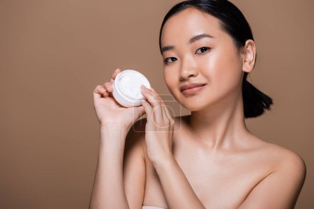 Téléchargez les photos : Pretty asian woman with naked shoulders holding container with cosmetic cream isolated on brown - en image libre de droit