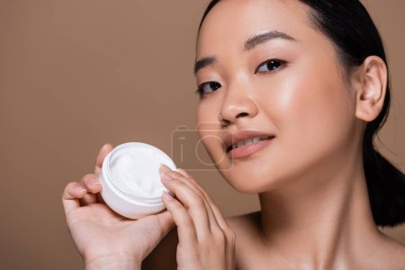 Téléchargez les photos : Young asian woman holding cosmetic cream and looking at camera isolated on brown - en image libre de droit