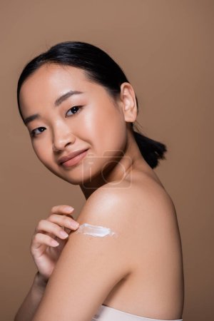 Smiling asian woman applying cosmetic cream on shoulder isolated on brown 