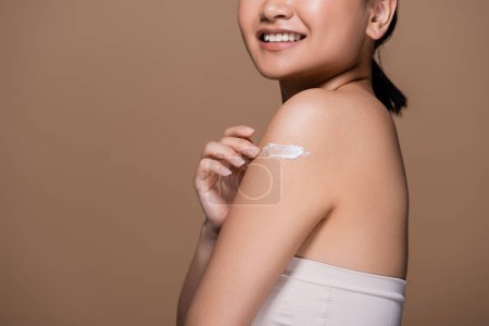 Cropped view of smiling woman in top applying cosmetic cream on shoulder isolated on brown 
