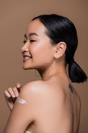 Cheerful asian model with naked shoulders applying cosmetic cream and looking away isolated on brown 