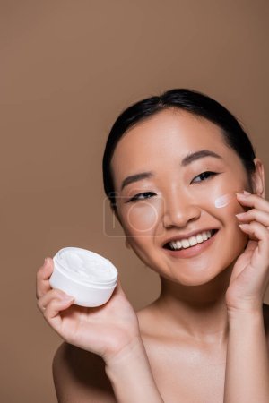Téléchargez les photos : Positive asian woman with naked shoulders applying face cream isolated on brown - en image libre de droit
