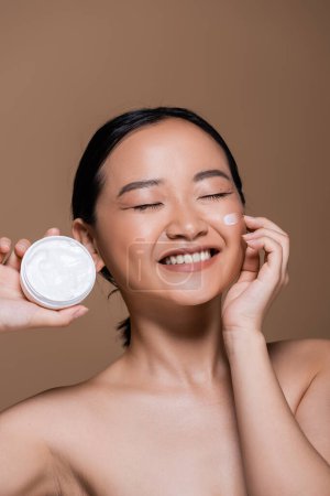 Pretty asian woman with naked shoulders holding cosmetic cream isolated on brown 
