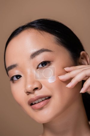 Portrait of young asian woman applying face cream on cheek isolated on brown 