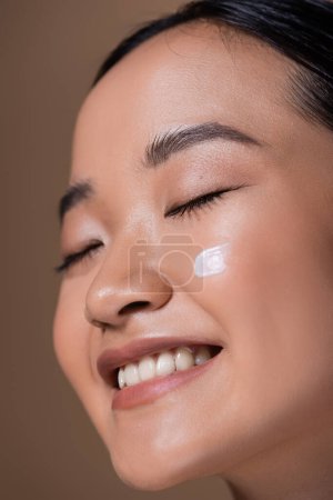 Photo for Close up view of positive asian woman with cream on cheek smiling isolated on brown - Royalty Free Image
