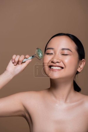 Smiling asian woman with naked shoulders holding jade roller isolated on brown 