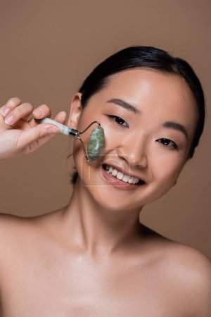 Cheerful asian woman with naked shoulders holding jade roller isolated on brown 