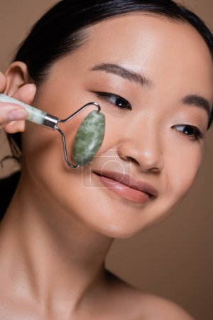 Portrait of asian woman holding jade roller near cheek isolated on brown 