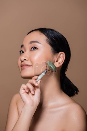 Young asian woman with naked shoulders holding jade roller and looking away isolated on brown 