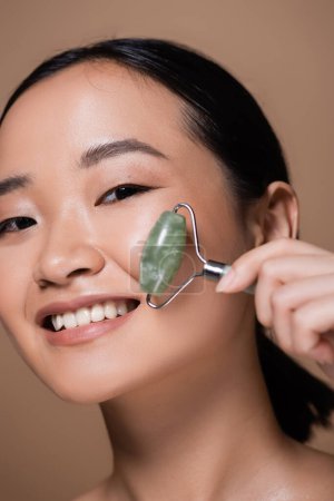 Portrait of cheerful asian woman holding jade roller and looking at camera isolated on brown 